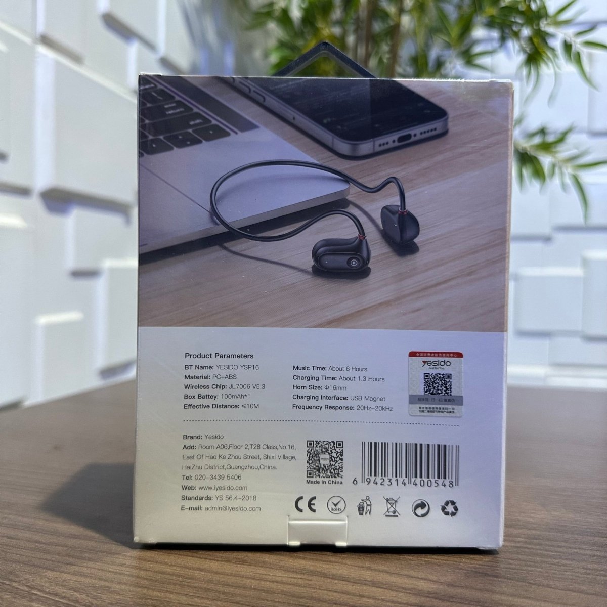 Product packaging with wireless earbuds on a desk, featuring a laptop and phone.