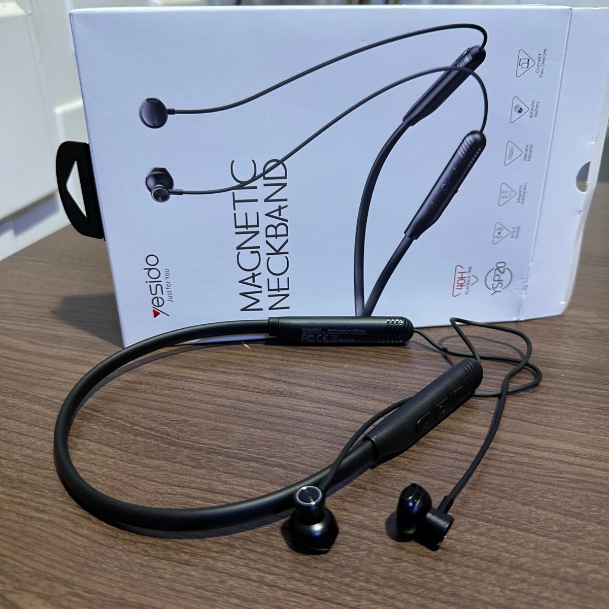 Black magnetic neckband with earpieces on a wooden surface in front of its packaging box.