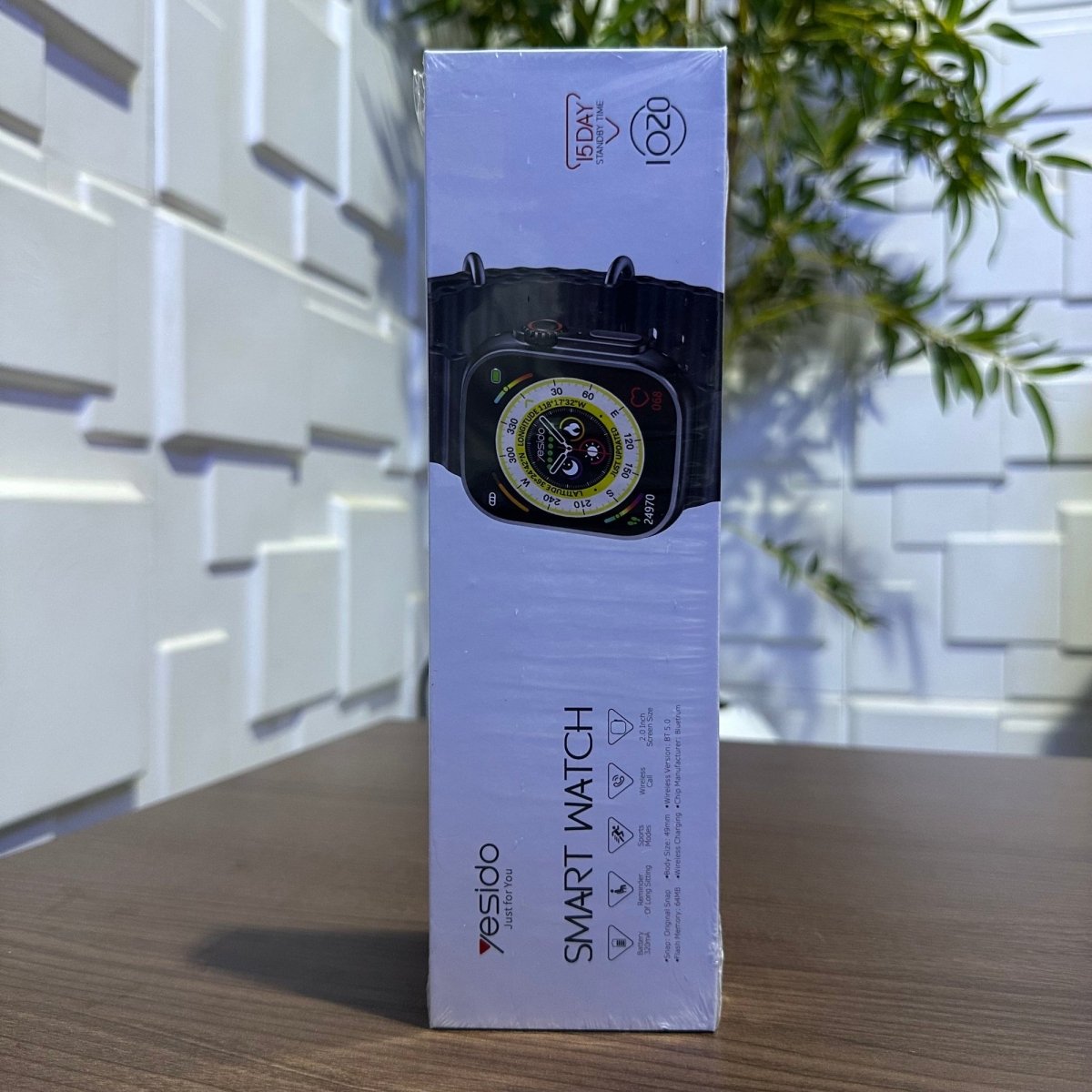Box of a smartwatch on a wooden surface with a light-colored wall and plant in the background