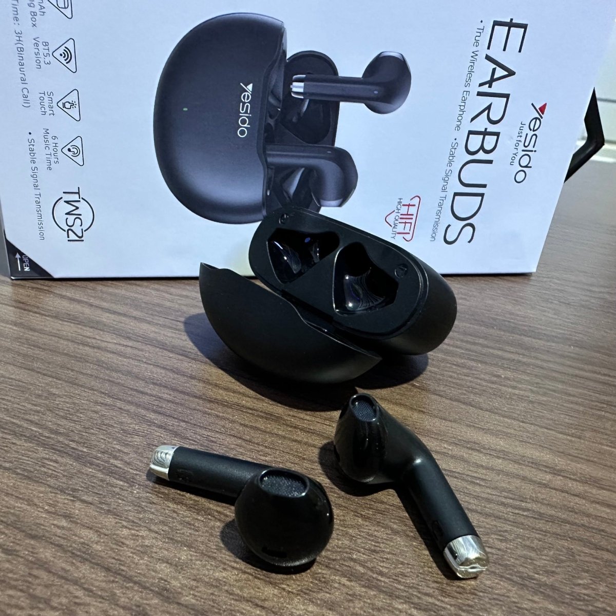 Black wireless earbuds with charging case on a wooden surface, brand 'Yesido' visible.