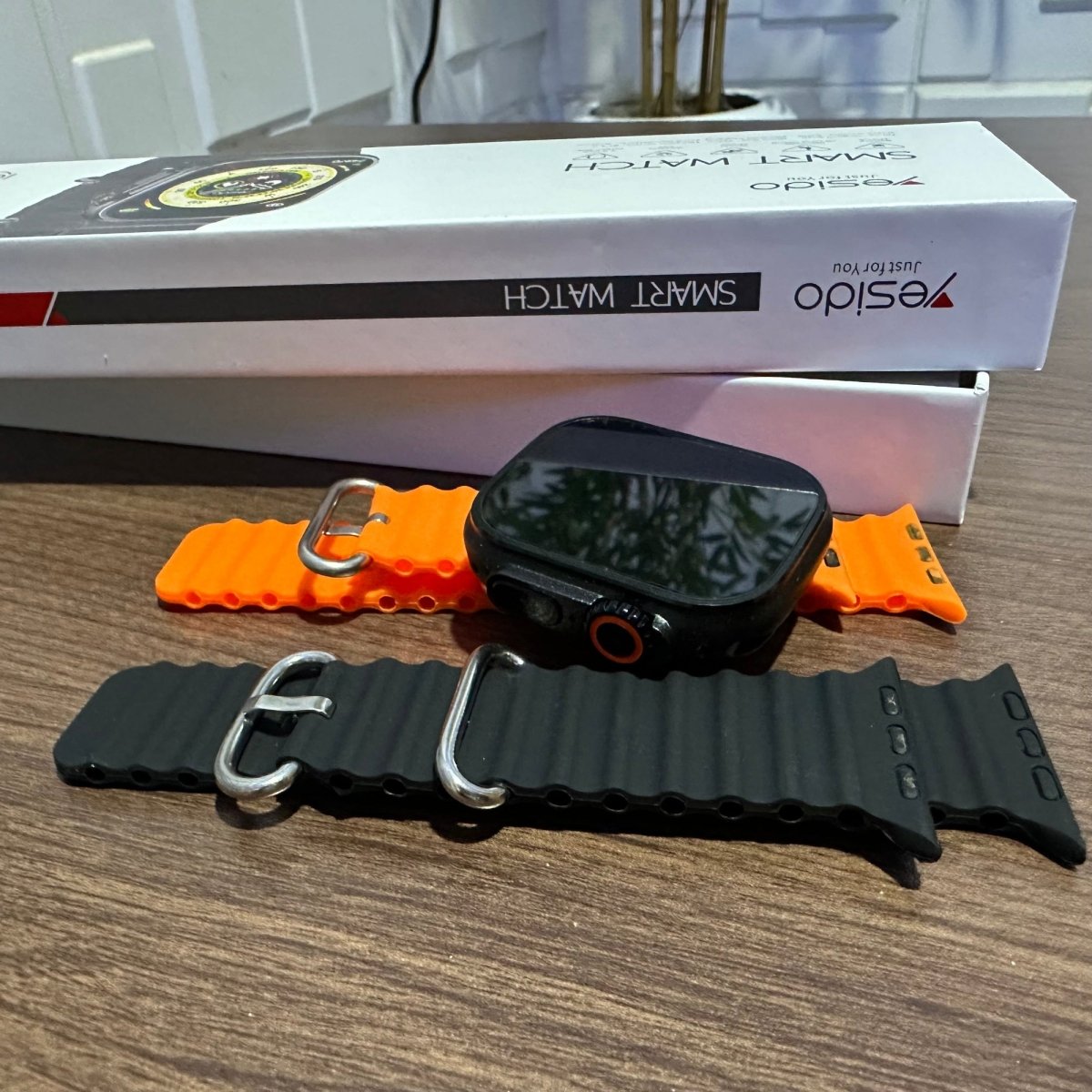 A black smartwatch with an orange strap is on a wooden surface, and a white box labeled 'YESIDO' is in the background.