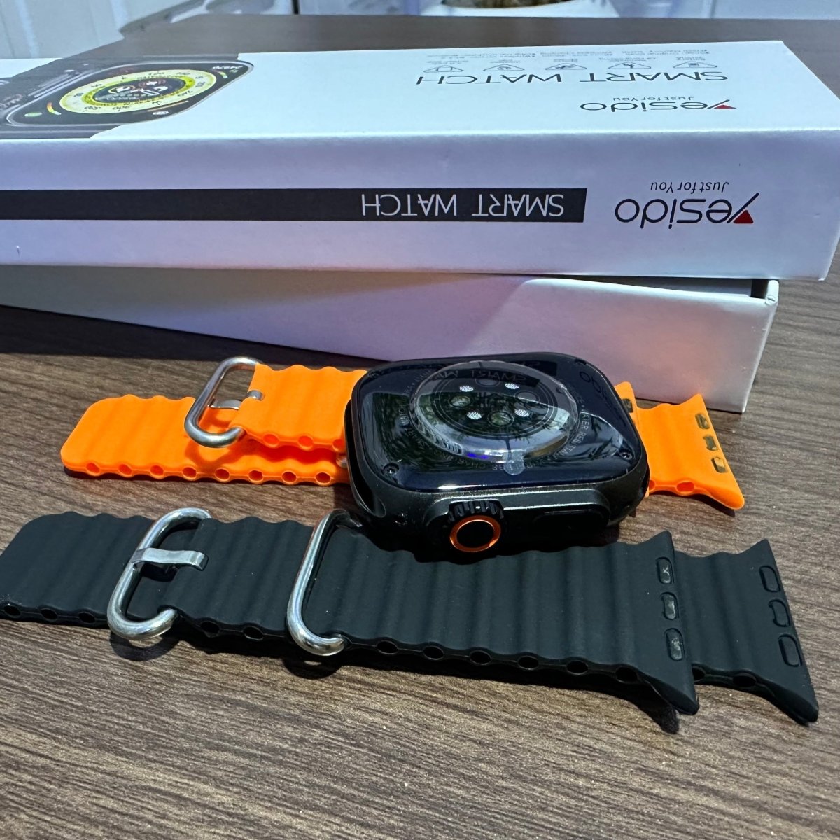 Smartwatch with black and orange straps on a table in front of a white box labeled 'Eseido'.